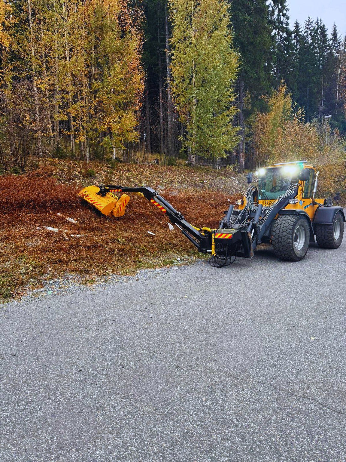 Kunnossapito ja koneurakointi -palvelun keltainen traktori raivaa pitkällä mekaanisella käsivarrellaan ruskeita lehtiä ja roskia tienvarsilta syksyn väristen puiden taustaa vasten. , JR Pihahuolto Oy Kuopio Kunnossapito ja koneurakointi -palvelun keltainen traktori raivaa pitkällä mekaanisella käsivarrellaan ruskeita lehtiä ja roskia tienvarsilta syksyn väristen puiden taustaa vasten. , JR Pihahuolto Oy Kuopio