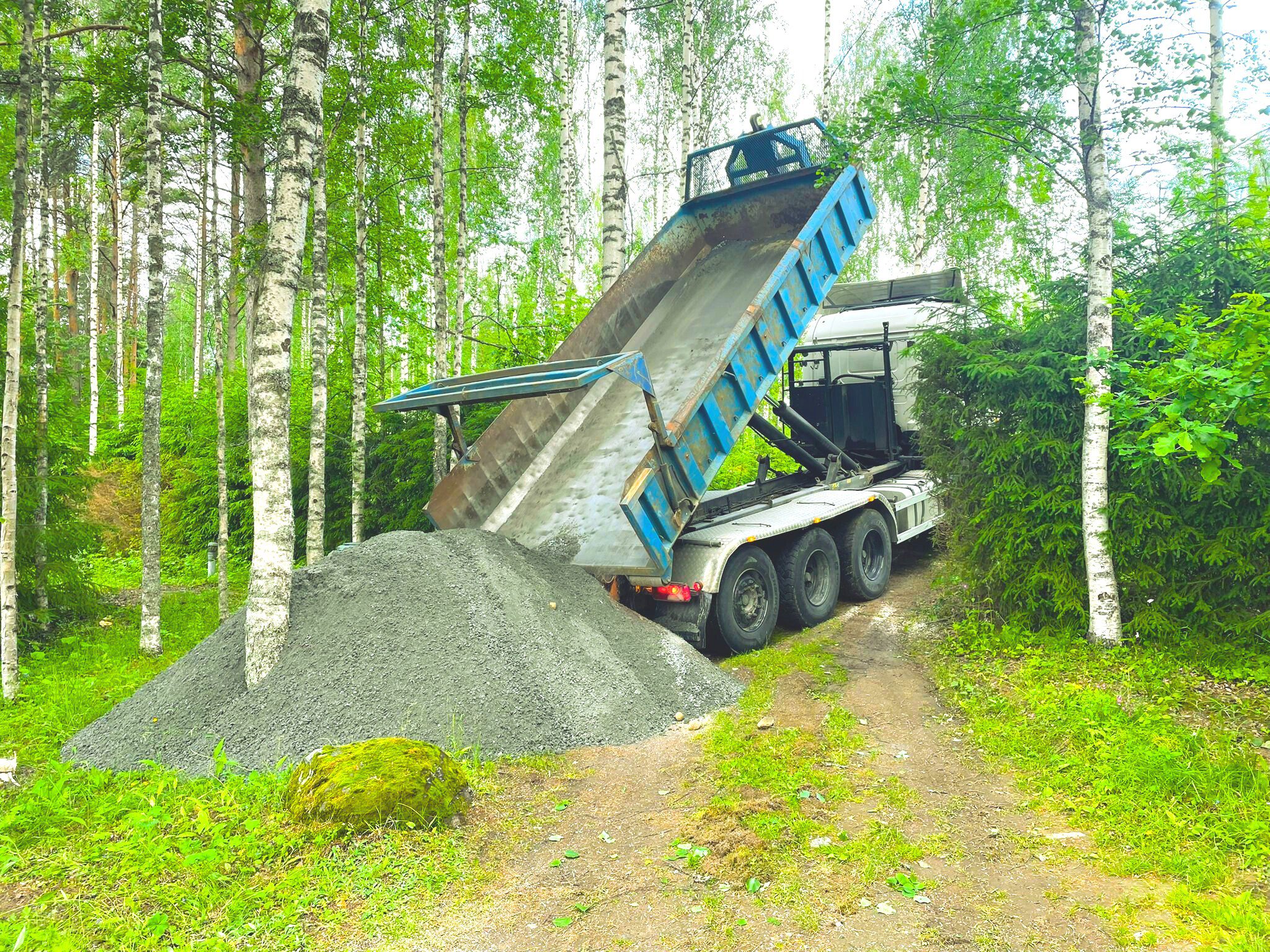 Jr pihahuollon kuorma-auto purkaa suuren sorakasan metsäpolulle, jota ympäröivät vihreät puut ja pensaat. Kuorma-auton lava on nostettu ylös, jolloin soraa valuu maahan rehevällä, metsäisellä alueella. | JR Pihahuolto Oy, Kuopio Jr pihahuollon kuorma-auto purkaa suuren sorakasan metsäpolulle, jota ympäröivät vihreät puut ja pensaat. Kuorma-auton lava on nostettu ylös, jolloin soraa valuu maahan rehevällä, metsäisellä alueella. | JR Pihahuolto Oy, Kuopio