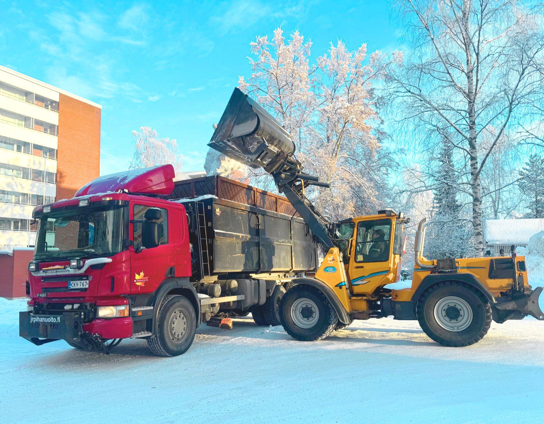 JR Pihahuollon keltainen kuormaaja kaataa lunta suureen punamustaan kuorma-autoon lumisella kadulla, jonka taustalla on korkeita puita ja kerrostalo kirkkaan sinisen taivaan alla. | JR Pihahuolto Oy, Kuopio JR Pihahuollon keltainen kuormaaja kaataa lunta suureen punamustaan kuorma-autoon lumisella kadulla, jonka taustalla on korkeita puita ja kerrostalo kirkkaan sinisen taivaan alla. | JR Pihahuolto Oy, Kuopio