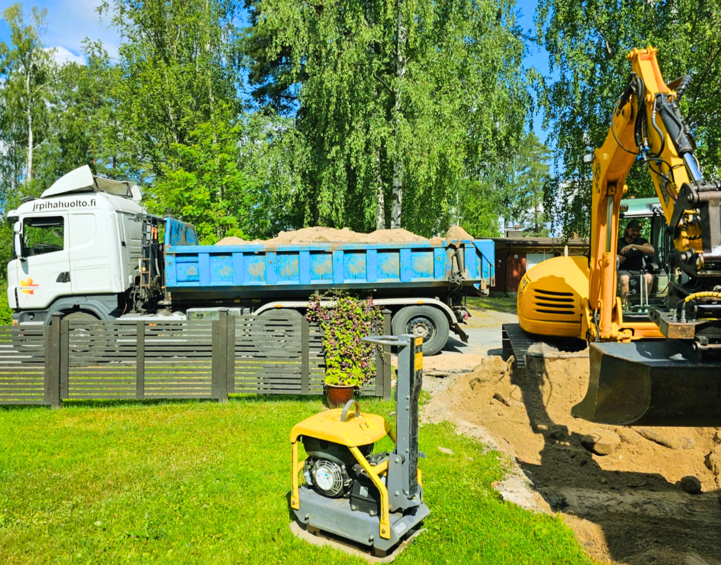 Valkoinen hiekalla täytetty kuorma-auto on pysäköity aidan lähelle, kun kaivinkone, joka on osa jr pihahuolto-hanketta, kaivaa ja siirtää hiekkaa. Nurmikolla istuu lautaspuristin ja ruukkukasvi, ja taustalla on puita ja rakennus. | JR Pihahuolto Oy, Kuopio Valkoinen hiekalla täytetty kuorma-auto on pysäköity aidan lähelle, kun kaivinkone, joka on osa jr pihahuolto-hanketta, kaivaa ja siirtää hiekkaa. Nurmikolla istuu lautaspuristin ja ruukkukasvi, ja taustalla on puita ja rakennus. | JR Pihahuolto Oy, Kuopio