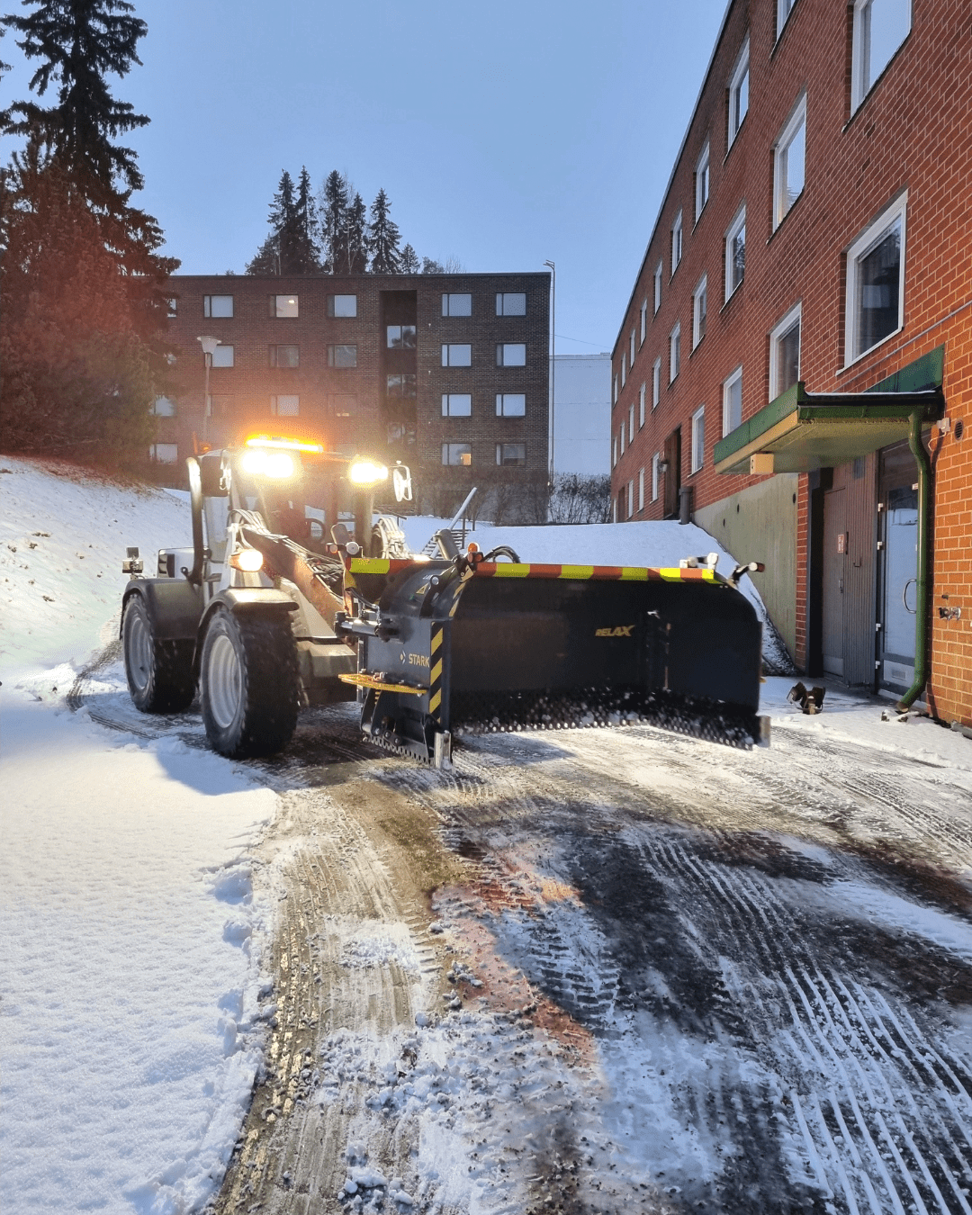 Traktori, jossa on kunnossapidon ja koneurakointipalveluihin kuuluva lumiaura, raivaa lumipeitteistä ajotietä punatiilisen kerrostalon vieressä iltahämärässä, valot ovat päällä ja taustalla näkyy puita ja toinen rakennus. , JR Pihahuolto Oy Kuopio Traktori, jossa on kunnossapidon ja koneurakointipalveluihin kuuluva lumiaura, raivaa lumipeitteistä ajotietä punatiilisen kerrostalon vieressä iltahämärässä, valot ovat päällä ja taustalla näkyy puita ja toinen rakennus. , JR Pihahuolto Oy Kuopio